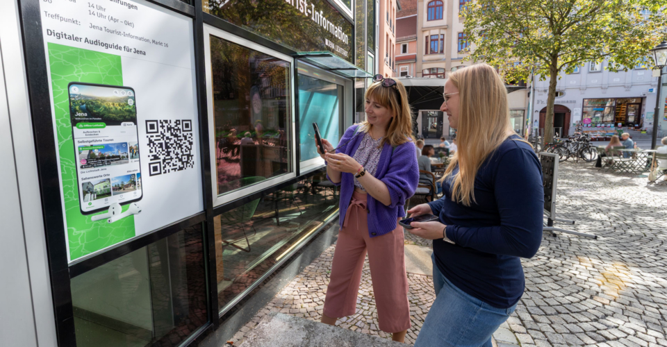 Zwei Frauen scannen vor der Jena Tourist-Information einen QR-Code auf einem Plakat für eine digitale Stadtführung.