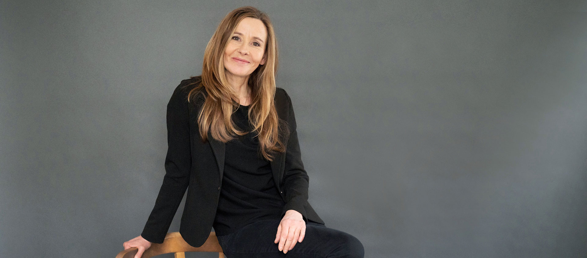 Andrea Wulf sitzt lächelnd auf einem Holzstuhl vor grauem Hintergrund, trägt schwarze Kleidung und schwarze Sneakers