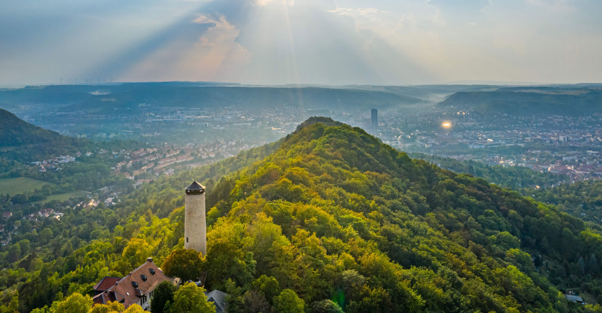 Fuchsturm auf bewaldetem Hügel mit Blick auf die Stadt Jena und Sonnenstrahlen durch Wolken am Himmel
