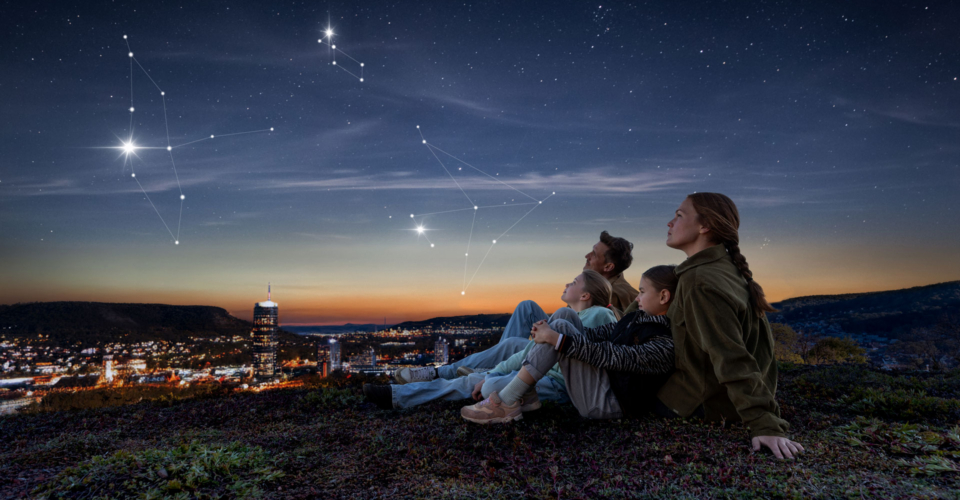 Familie sitzt auf einer Anhöhe über Jena und blickt in den Abendhimmel mit hervorgehobenen Sternbildern