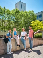 Vier Studierende stehen vor einer Mauer auf dem Campus der Ernst-Abbe-Hochschule Jena mit einem hohen Turm und Bäumen im Hintergrund.
